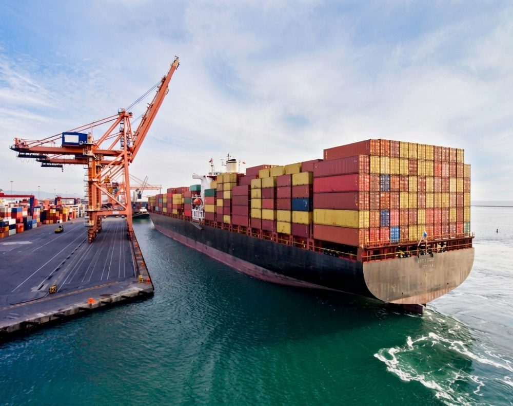 Cargo ship docked at a port, loaded with containers, near an orange crane and blue water.