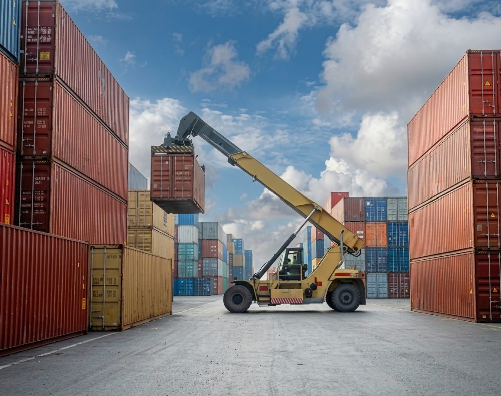 Forklift lifting a shipping container in a busy port with many colorful containers stacked high under a cloudy sky.