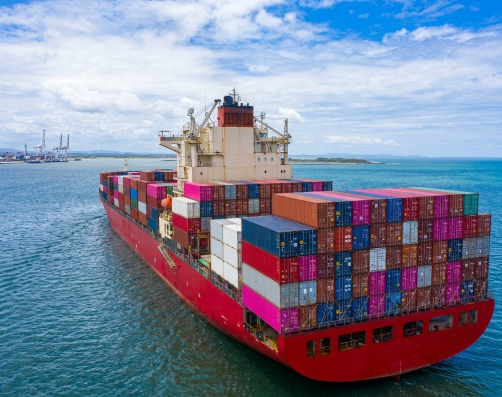 Red cargo ship sailing in ocean, carrying stacks of shipping containers.