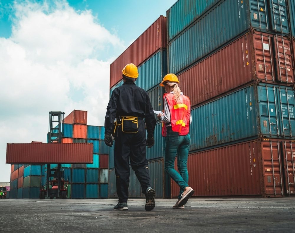 Two people in safety vests and hard hats at a shipping port, surrounded by cargo containers.