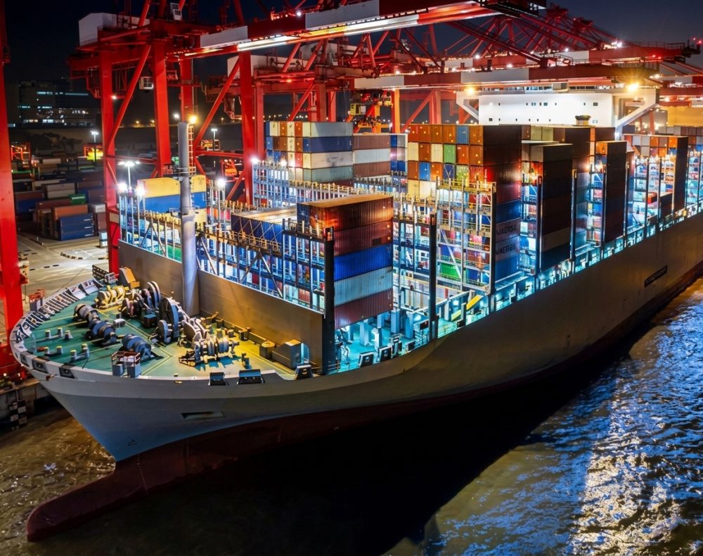 Cargo ship at a dock, illuminated at night with red cranes loading shipping containers.