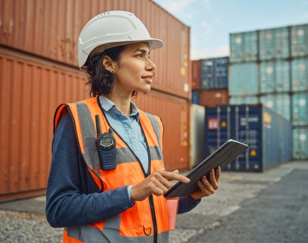 Woman in hard hat and safety vest, holding tablet, surveys shipping containers.